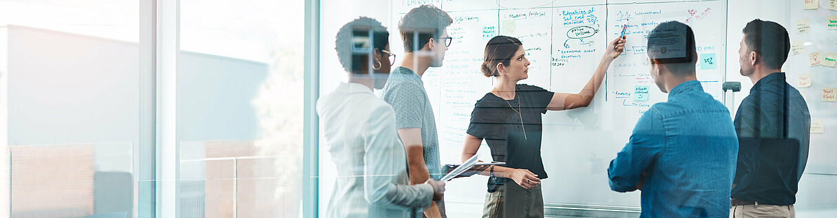 Teammeeting in einem modernen Büro: Mitarbeitende analysieren Aufgaben auf einem Whiteboard in einem agilen Arbeitsumfeld mit Fokus auf Projektplanung und Zusammenarbeit.